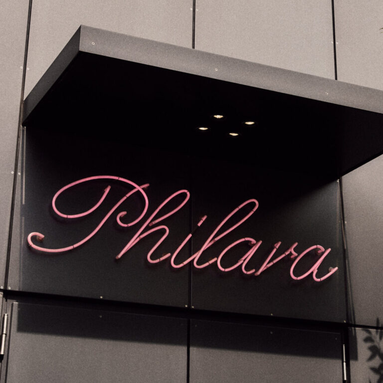 Ein rosafarbenes Neonschild mit der Aufschrift Philara an einem modernen grauen Gebäude, mit Baumschatten an der Wand, spiegelt das Engagement des Lokals für Nachhaltigkeit im Catering wider.