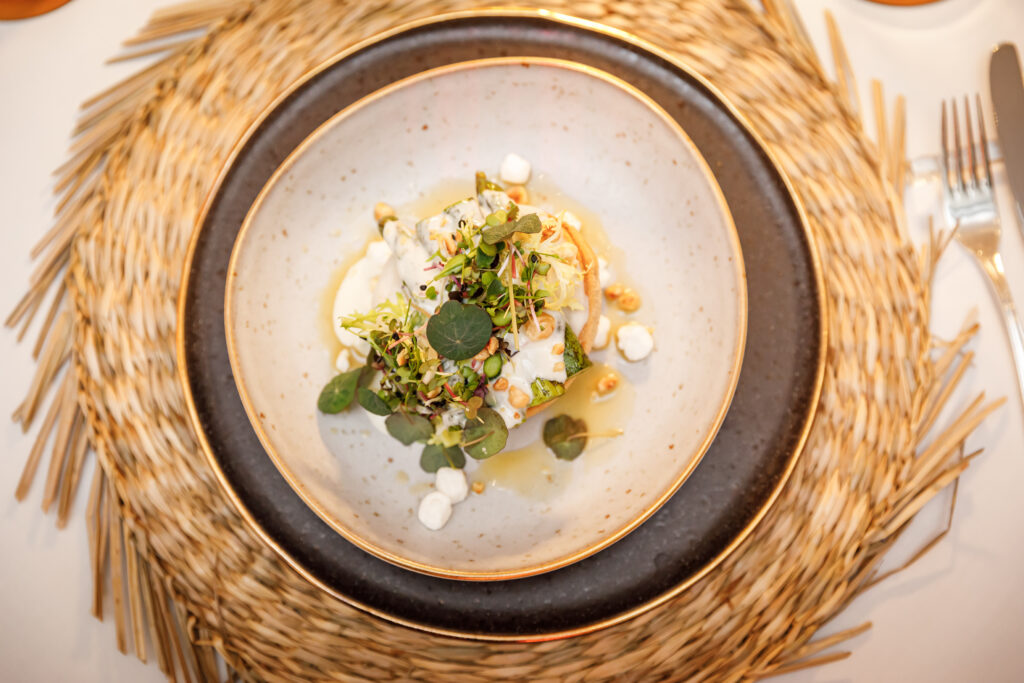 Ein Feinschmeckergericht mit Grünzeug und Soße, das in einer Schale auf einem geflochtenen Tischset serviert wird, neben dem das Besteck liegt.