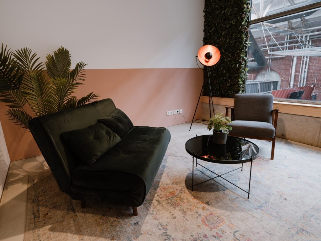 Moderne Eventlocation im Wohnzimmerstil mit einem grünen Sofa, einem Sessel, einem runden Couchtisch, einer Pflanze und einer Stehlampe an einem großen Fenster.