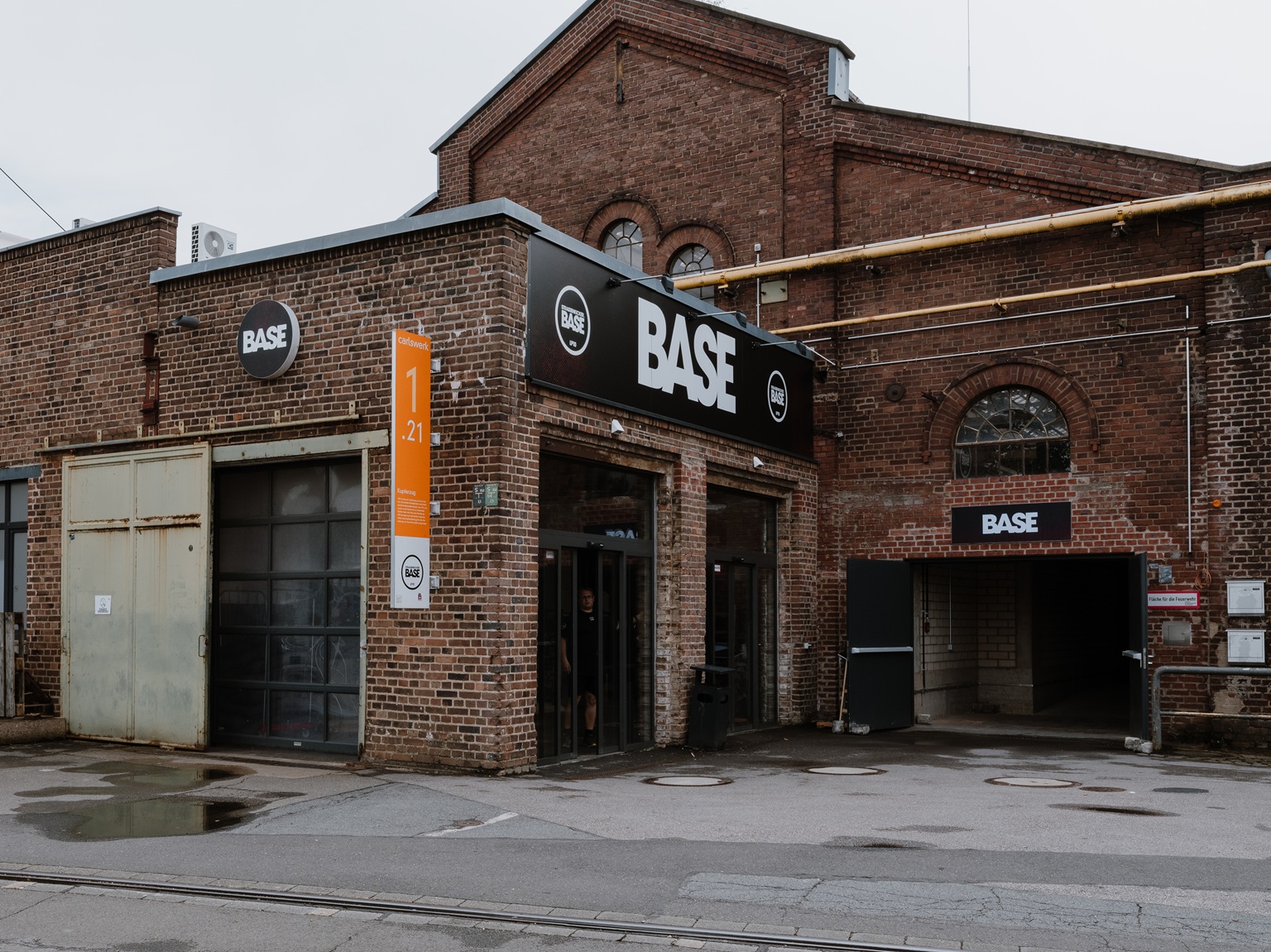 Industriegebäude aus Backstein mit BASE-Schildern über den Eingängen und Garagentoren an einem bewölkten Tag.
