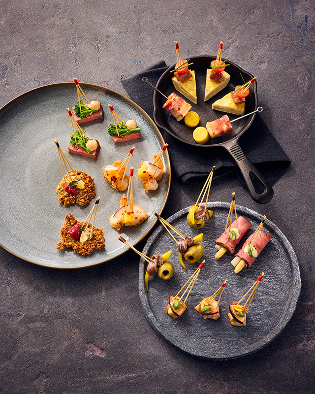 Verschiedene Vorspeisenspieße auf Tellern und einer kleinen Pfanne, ordentlich auf einer strukturierten Oberfläche angeordnet - perfekte Häppchen zum Genießen, bevor man Philara 14 Places erkundet.