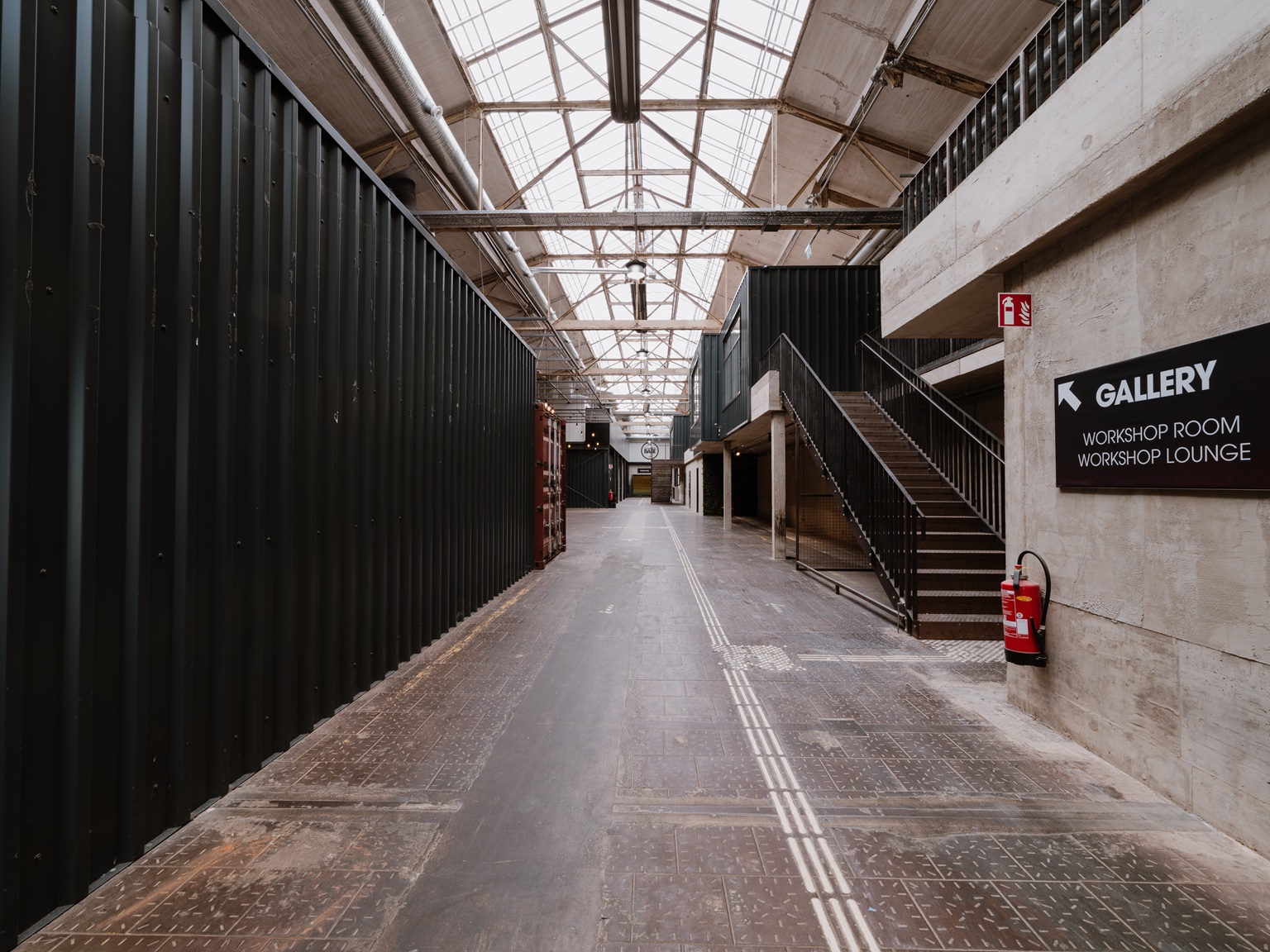 Industriehalle mit Oberlichtern, Metallwänden, Treppen und einem Schild, das auf eine Galerie und Werkstatträume hinweist.