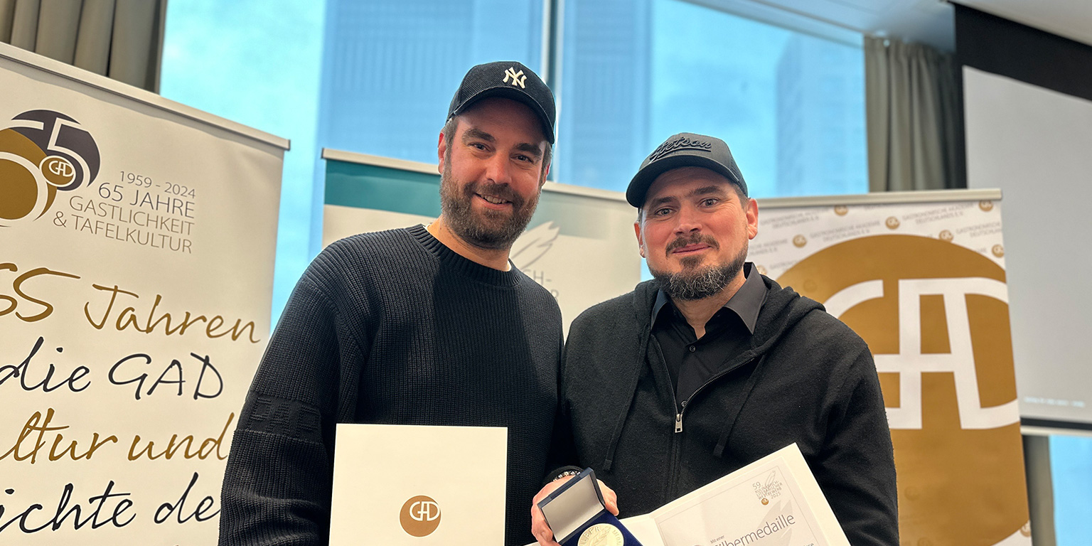 Zwei Männer mit Mützen halten Urkunden und eine Medaille in der Hand. Sie stehen zusammen bei einer Veranstaltung zur Verleihung der Philara 14 Places.