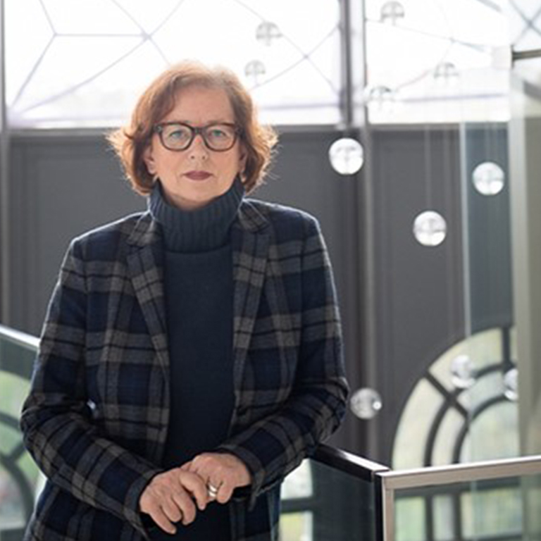 Eine Frau mit Brille und kurzen Haaren in einer karierten Jacke steht in einem Innenraum an einem Glasgeländer und blickt in die Kamera - ein Zeichen für ihr Engagement für Nachhaltigkeit im Catering.