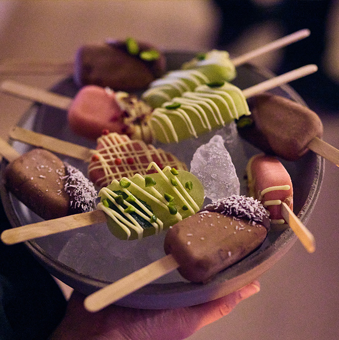 Eine Schale mit verschiedenen Minieis am Stiel, umhüllt von Schokolade und bunten Toppings, serviert auf Eis - ein süßes Highlight, das durch umweltfreundliche Präsentation und Zutaten Nachhaltigkeit im Catering zeigt.