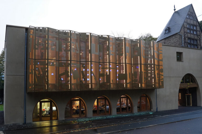 Ein modernes Gebäude mit Rundbogenfenstern und einer gemusterten, halbtransparenten Fassade steht neben dem historischen Kirberg Catering Sachsenturm Köln, einem älteren Bau mit steilem Dach.