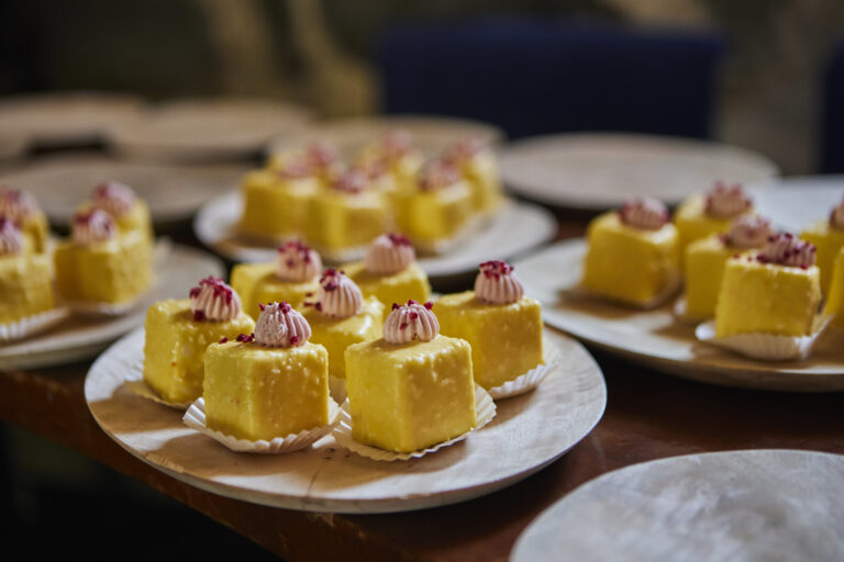 Mehrere Teller mit gelben, quadratischen Kuchen mit rosa-weißem Zuckerguss stehen auf einem Tisch und zeigen die elegante Präsentation von Kirberg Catering im Sachsenturm Köln.
