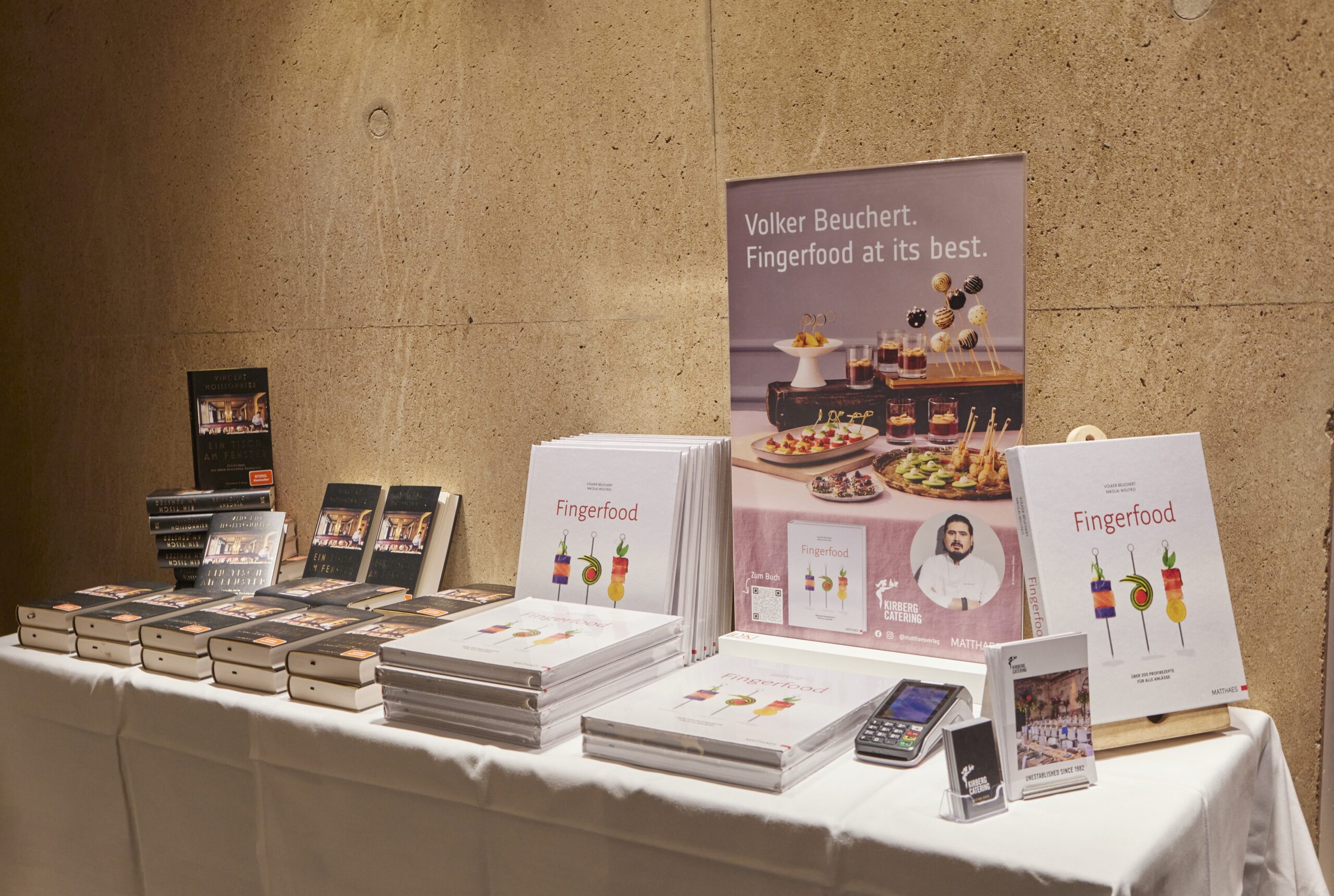 Tischaufsteller mit Stapeln von Fingerfood-Büchern, Plakaten von Kirberg Catering Sachsenturm Köln und einem Kartenzahlungsautomaten vor einer beigen Wand.